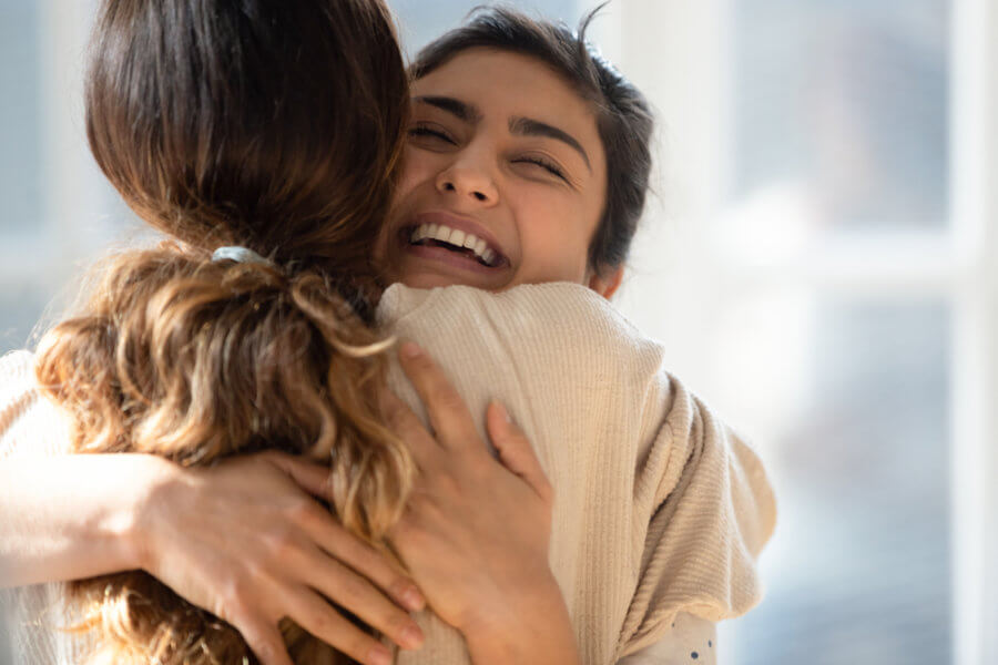 Two women smiling and hugging each other; how social factors influence your addiction